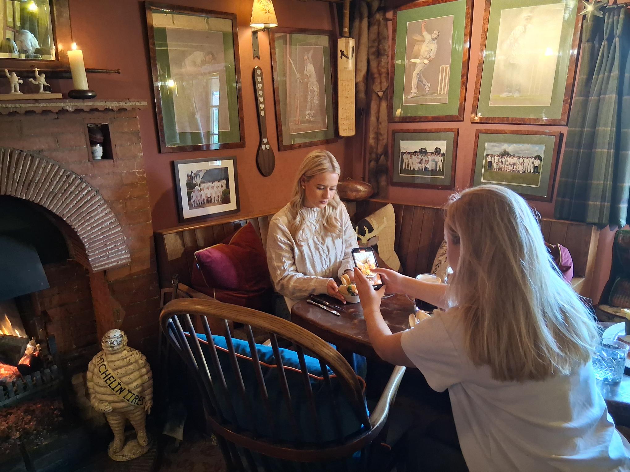 A woman is taking a photo of another woman who is holding a bowl of food in a country pub interior. 