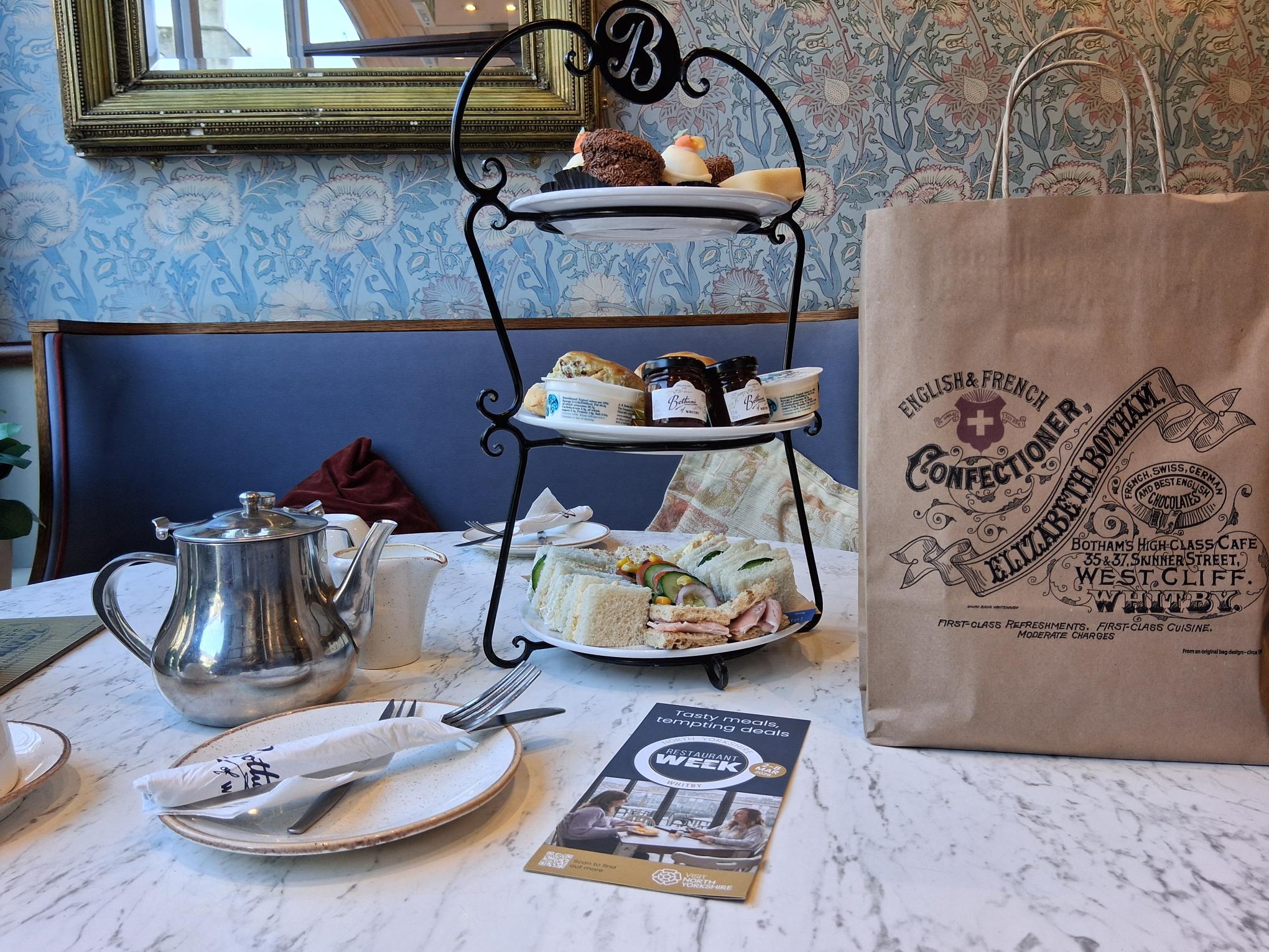 A three tiered afternoon tea cake stand filled with sandwiches and cakes, alongside a pot of tea, with a Botham's of Whitby branded paper bag.