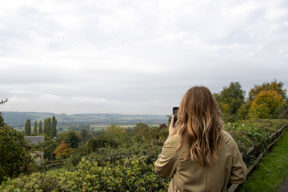 A woman take a photo on her phone of trees and hills in the distance