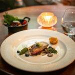 a delicately arranged starter on a white plate rests on a round wooden table,