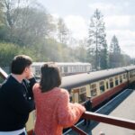 A young couple look out over a steam train in a station from the footbridge