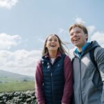 A young couple are standing in front of a dry stone wall on a sunny day, they smiling as they look at the view, behind them are the rolling hills of the Yorkshire dales.