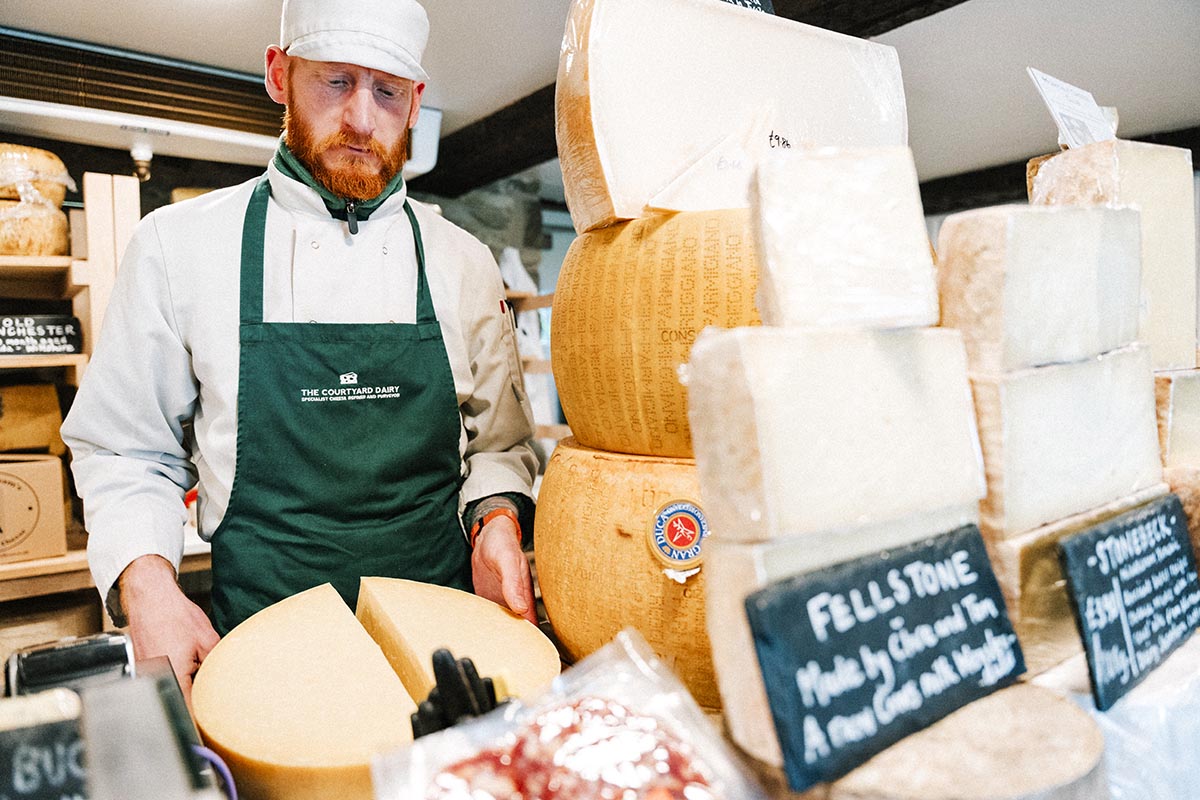A man in a cheese shop is cutting a large cheese in half, his is surrounded but many other cheeses. In the foreground are name labels for some of the varieties, "Stonebeck" and "Fellstone"