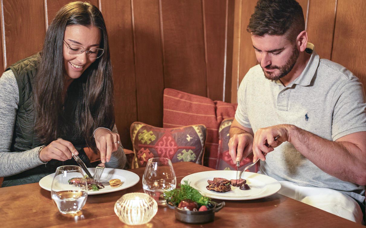 A couple sitting at a corner table against a wood panel backdrop into a meal tuck into elegent starters