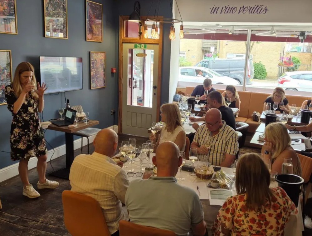 A room of people are seated at tables, with glasses of wine, while a sommelier stands in front of them describing the wine they are tasting